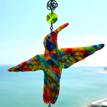 A handmade hummingbird suncatcher crafted from colorful polymer clay, featuring a detailed hummingbird design in shades of green, blue, and pink, suspended from a gold chain with a faceted blue maple leaf glass crystal that casts rainbows in the sunlight. Perfect for nature lovers and whimsical window decor.