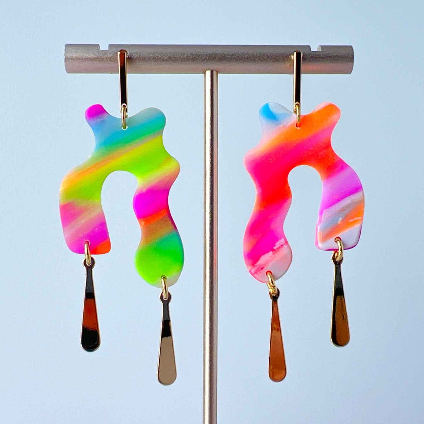 A pair of handmade neon abstract-shaped polymer clay earrings featuring rainbow marbled colors (green, pink, orange, purple, and coral) with shiny gold-plated vertical bar dangles. 