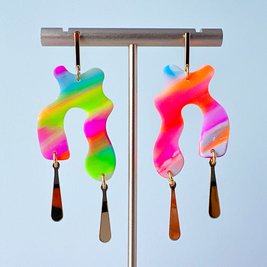 A pair of handmade neon abstract-shaped polymer clay earrings featuring rainbow marbled colors (green, pink, orange, purple, and coral) with shiny gold-plated vertical bar dangles. 