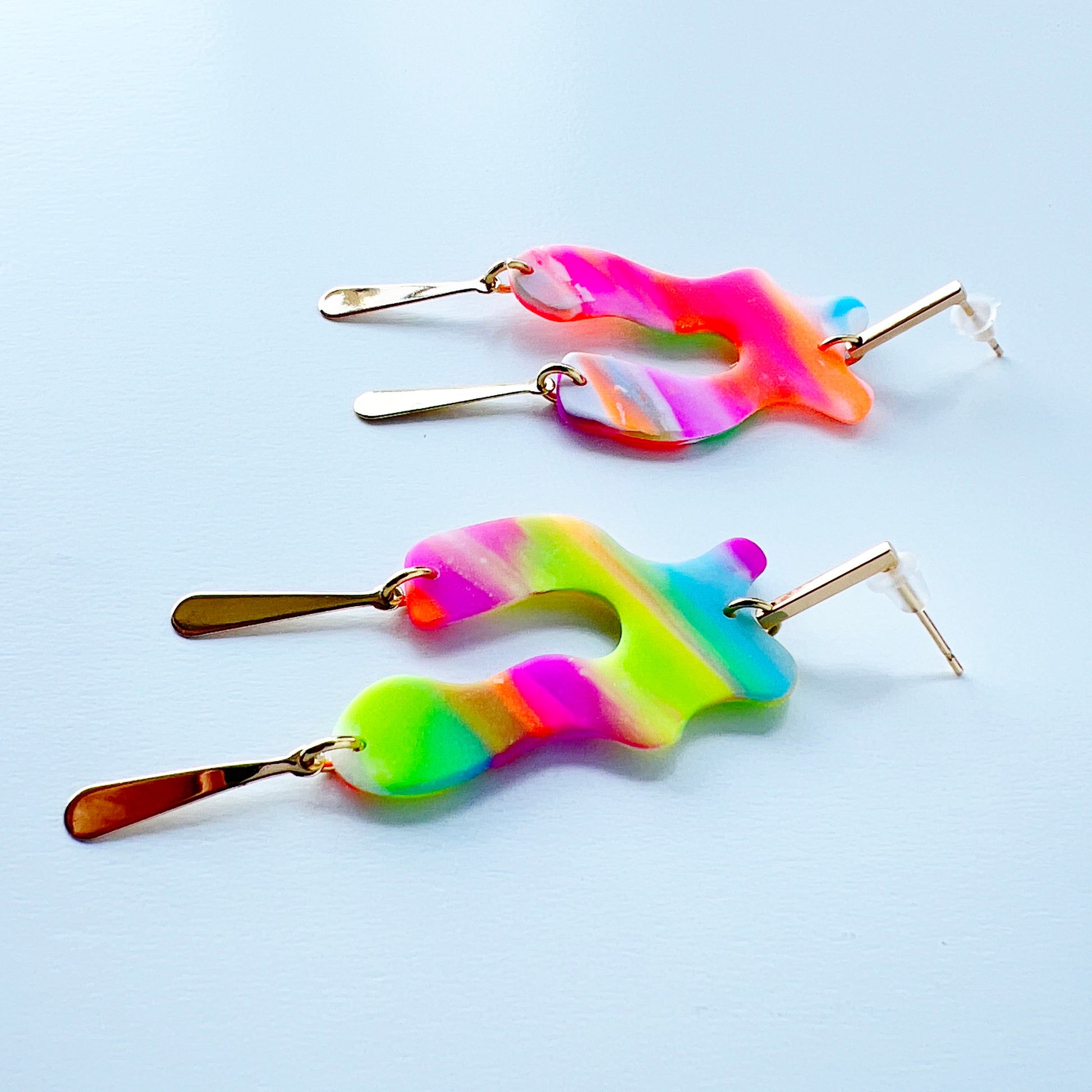 A pair of handmade neon abstract-shaped polymer clay earrings featuring rainbow marbled colors (green, pink, orange, purple, and coral) with shiny gold-plated vertical bar dangles.