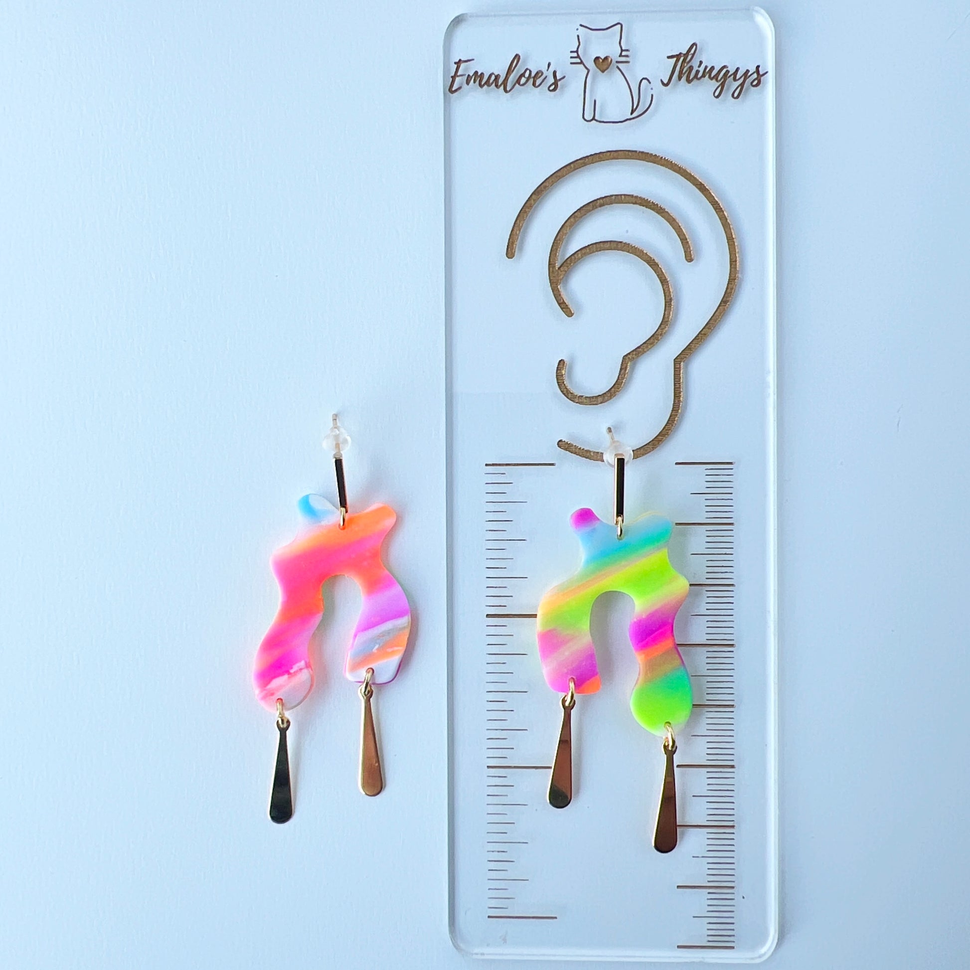 A pair of handmade neon abstract-shaped polymer clay earrings featuring rainbow marbled colors (green, pink, orange, purple, and coral) with shiny gold-plated vertical bar dangles. The pair is displayed on a branded ruler showing the length to be about 2.25 inches long