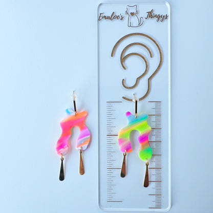 A pair of handmade neon abstract-shaped polymer clay earrings featuring rainbow marbled colors (green, pink, orange, purple, and coral) with shiny gold-plated vertical bar dangles. The pair is displayed on a branded ruler showing the length to be about 2.25 inches long