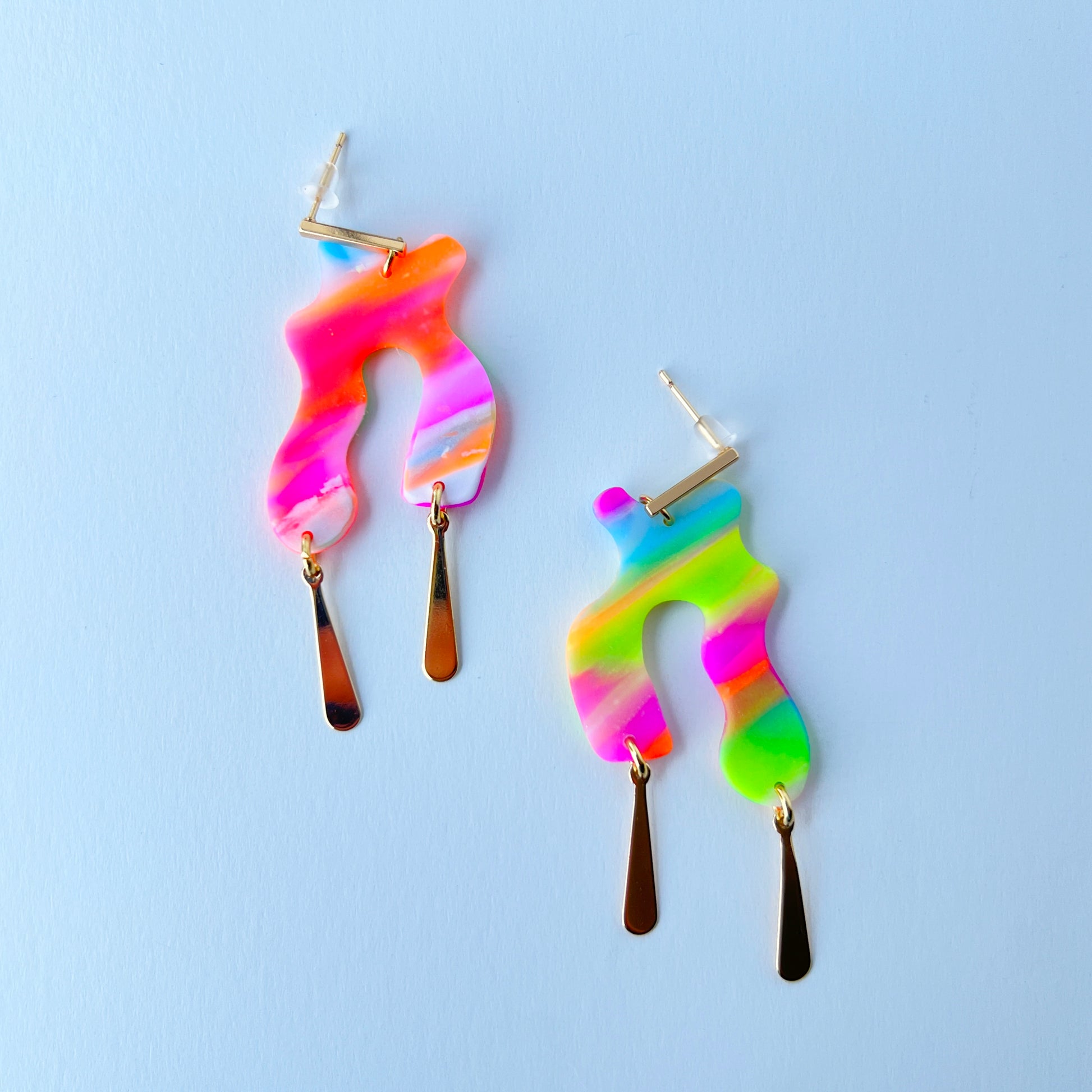 A pair of handmade neon abstract-shaped polymer clay earrings featuring rainbow marbled colors (green, pink, orange, purple, and coral) with shiny gold-plated vertical bar dangles.