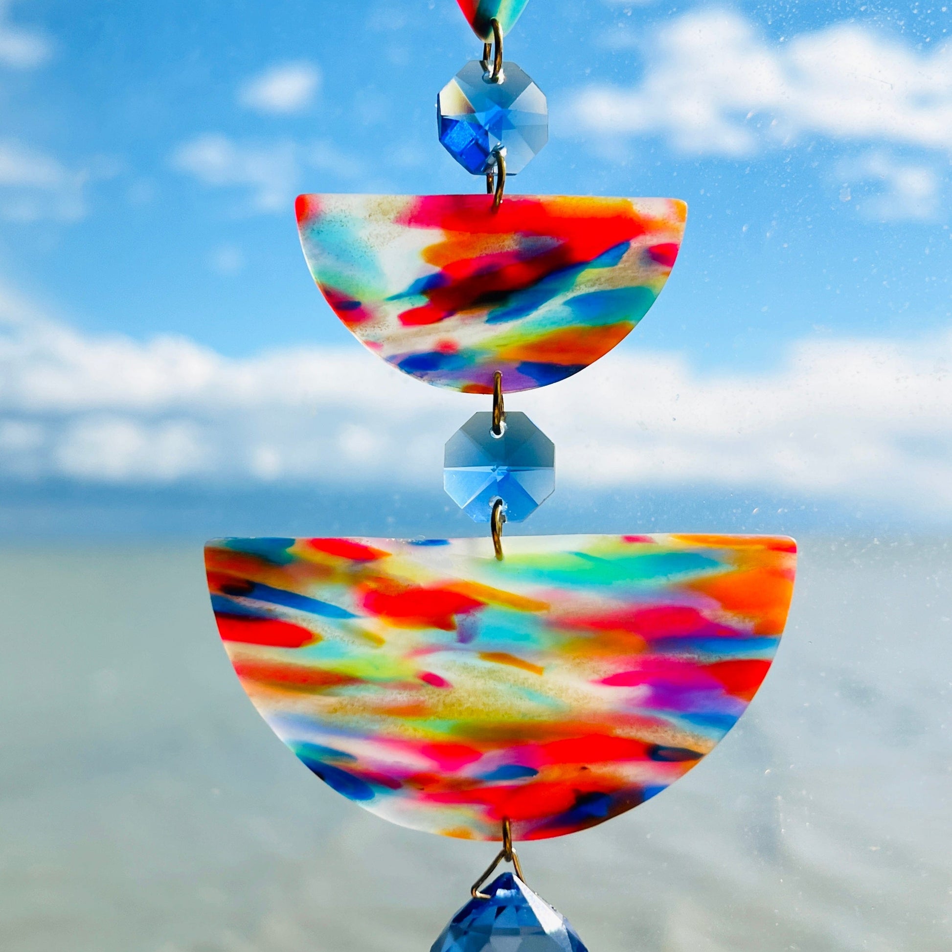 Paint Palette: Colorful handmade suncatcher with bold primary-toned polymer clay pieces in teardrop and arch shapes, paired with translucent blue faceted crystals and a rainbow chain.