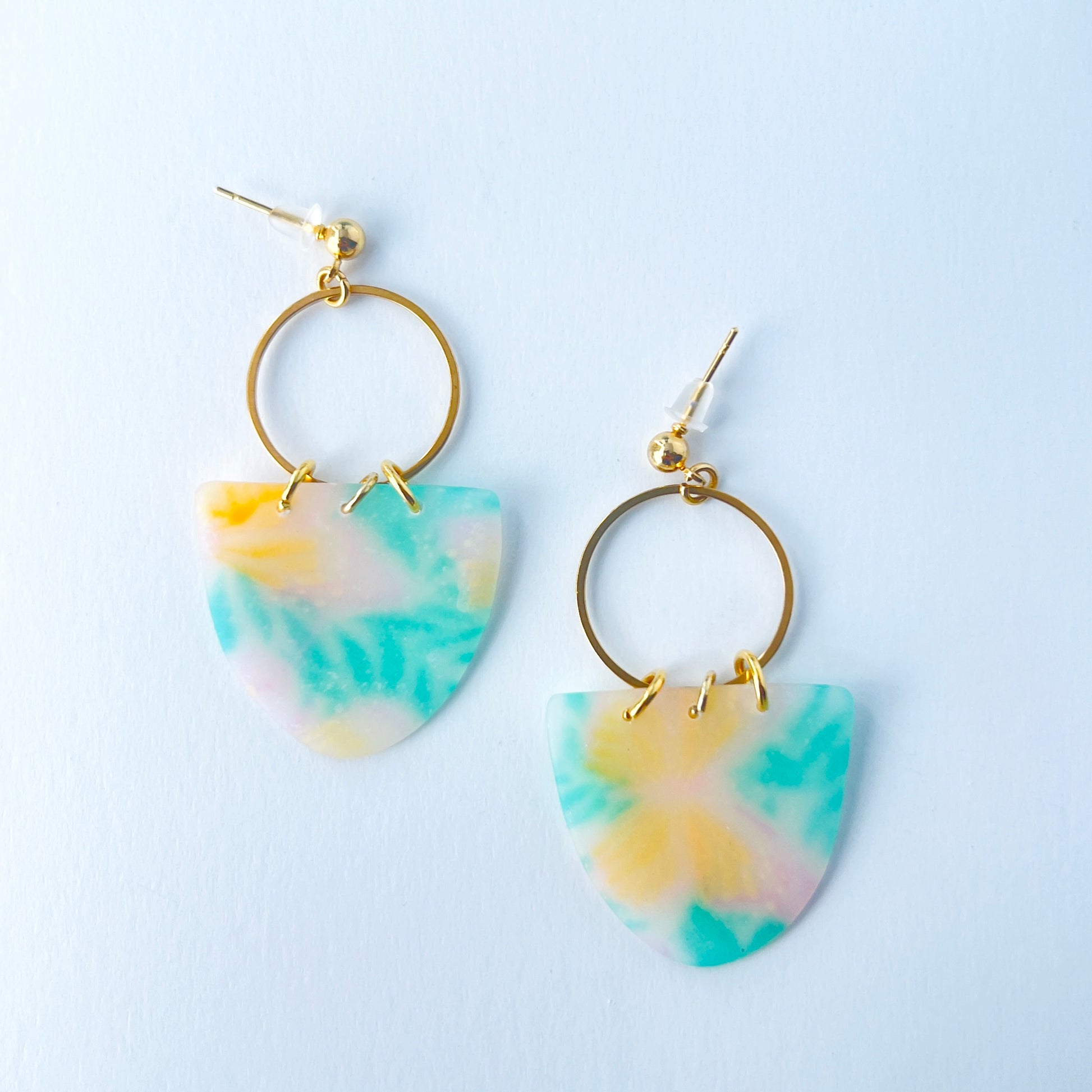 A pair of handmade earrings featuring semi-transparent polymer clay shields with soft pastel patterns of yellow butterflies and green leaves, attached to gold-plated hoops and ball posts.