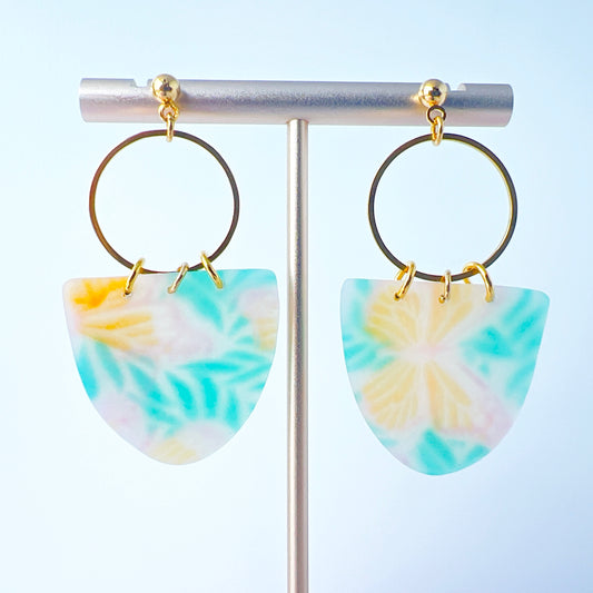 A pair of handmade earrings featuring semi-transparent polymer clay shields with soft pastel patterns of yellow butterflies and green leaves, attached to gold-plated hoops and ball posts.