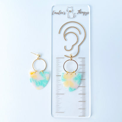 A pair of handmade earrings featuring semi-transparent polymer clay shields with soft pastel patterns of yellow butterflies and green leaves, attached to gold-plated hoops and ball posts.  They are displayed on a branded ruler showing the length of the earrings to be 2.25 inchs