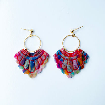 A pair of handmade fan-shaped polymer clay earrings displayed on a white background. The earrings feature gold-plated ball posts and hoops, with semi-transparent rainbow-colored clay and red etched fan-like lines. The marbling effect shows swirls of pink, blue, green, orange, and white.
