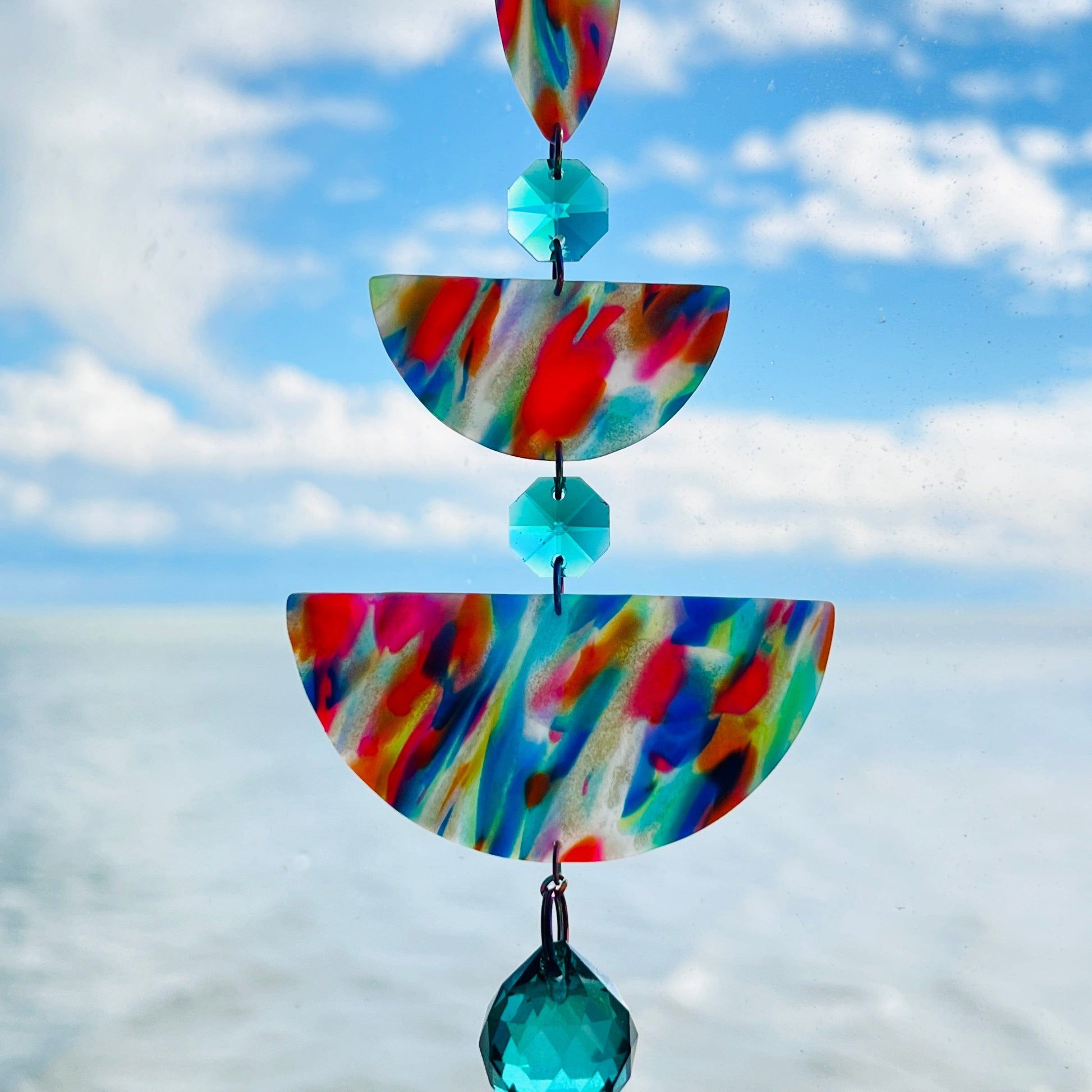 Rainbow Rain: A handmade suncatcher hanging in front of a blue sky, featuring marbled rainbow polymer clay shapes in teardrop and half-circle forms, accented with sea green glass crystals and a rainbow chain.