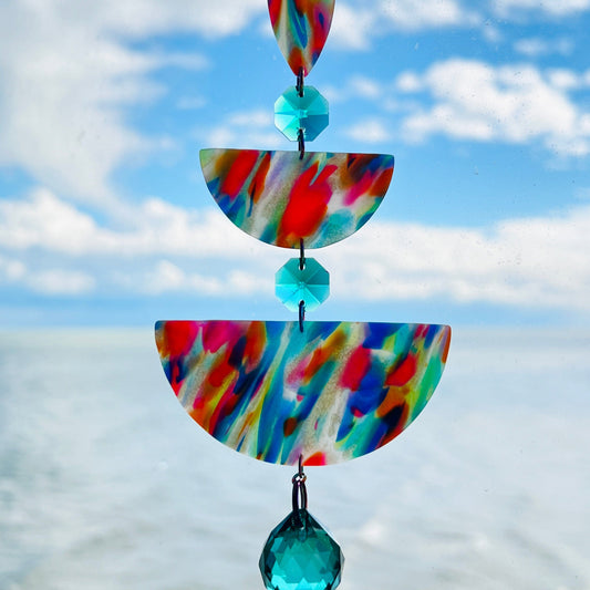 Rainbow Rain: A handmade suncatcher hanging in front of a blue sky, featuring marbled rainbow polymer clay shapes in teardrop and half-circle forms, accented with sea green glass crystals and a rainbow chain.