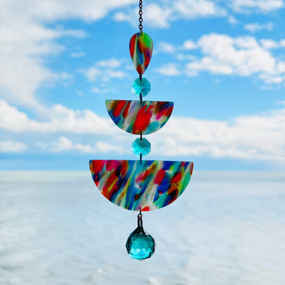 A handmade suncatcher hanging in front of a blue sky, featuring marbled rainbow polymer clay shapes in teardrop and half-circle forms, accented with sea green glass crystals and a rainbow chain.