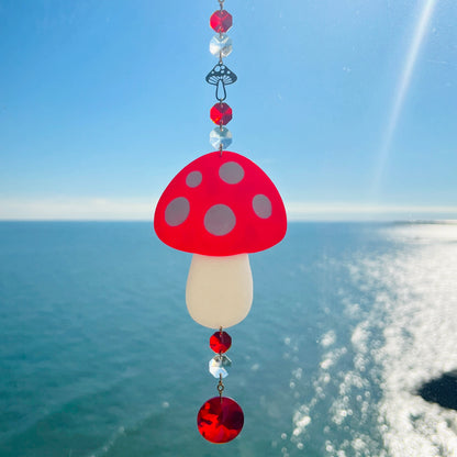 A whimsical handmade mushroom suncatcher crafted from polymer clay, featuring a red and white spotted toadstool design suspended from a gold chain, with a dangling faceted glass crystal that casts rainbows in the sunlight. Ideal for woodland, fairycore, or cottagecore decor.