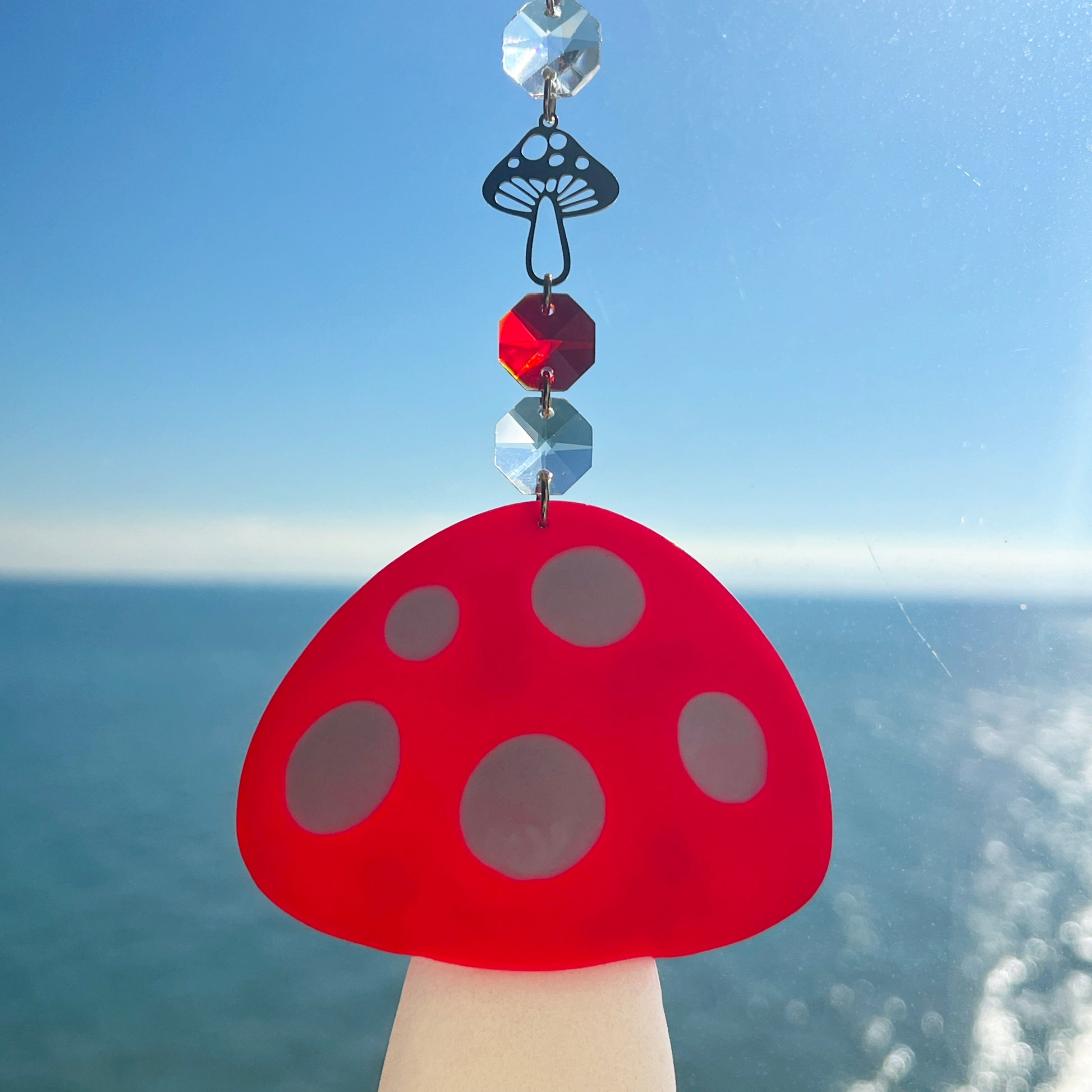 A whimsical handmade mushroom suncatcher crafted from polymer clay, featuring a red and white spotted toadstool design suspended from a gold chain, with a dangling faceted glass crystal that casts rainbows in the sunlight. Ideal for woodland, fairycore, or cottagecore decor.