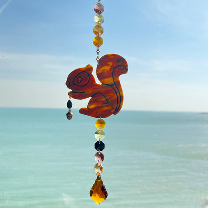 A handmade squirrel suncatcher featuring a brown polymer clay squirrel with a bushy tail and warm autumn tones, hanging from a gold chain with an amber faceted glass maple leaf crystal that reflects rainbow light in the sun. Ideal for woodland, fall, or forest-themed decor.