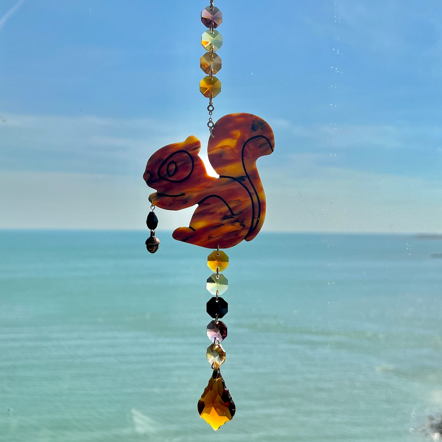 A handmade squirrel suncatcher featuring a brown polymer clay squirrel with a bushy tail and warm autumn tones, hanging from a gold chain with an amber faceted glass maple leaf crystal that reflects rainbow light in the sun. Ideal for woodland, fall, or forest-themed decor.