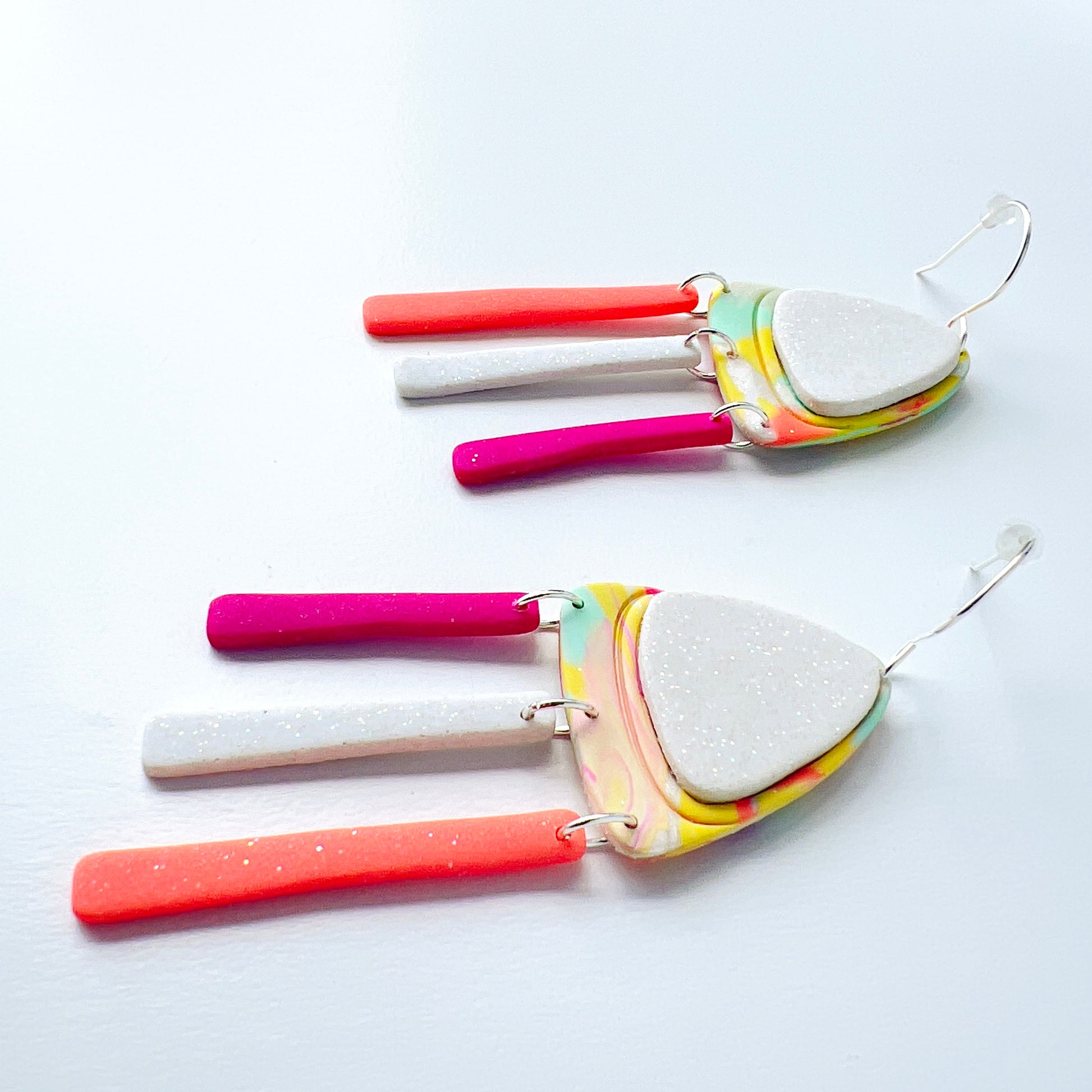 A pair of handmade polymer clay earrings with layered triangle shapes. The top layer is speckled white, sitting on a colorful marbled triangle of yellow, green, pink, and coral. Each earring features three dangles: one neon coral, one speckled white, and one bright pink, all hanging from silver stainless steel earwires.