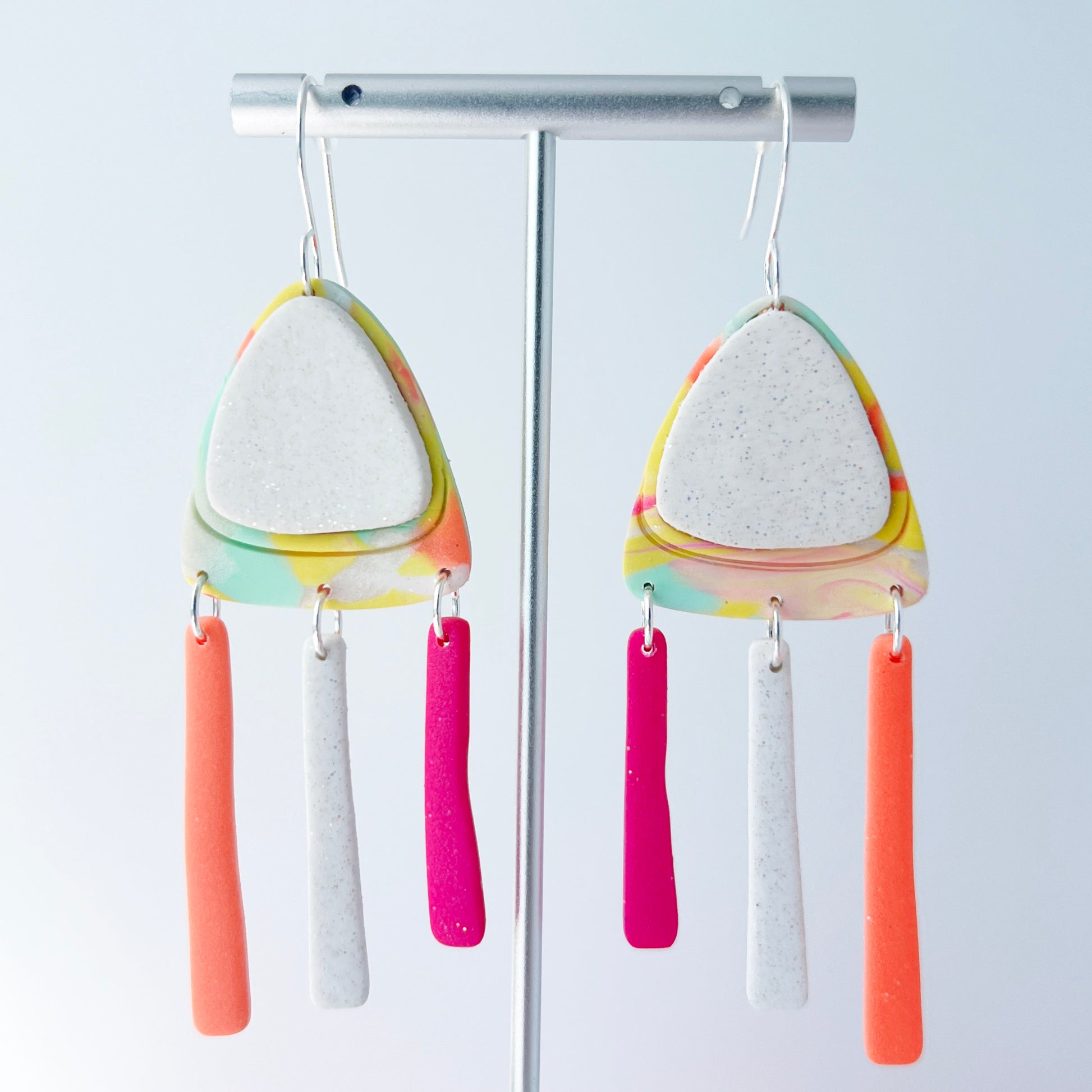A pair of handmade polymer clay earrings with layered triangle shapes. The top layer is speckled white, sitting on a colorful marbled triangle of yellow, green, pink, and coral. Each earring features three dangles: one neon coral, one speckled white, and one bright pink, all hanging from silver stainless steel earwires.
