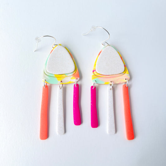 A pair of handmade polymer clay earrings with layered triangle shapes. The top layer is speckled white, sitting on a colorful marbled triangle of yellow, green, pink, and coral. Each earring features three dangles: one neon coral, one speckled white, and one bright pink, all hanging from silver stainless steel earwires.