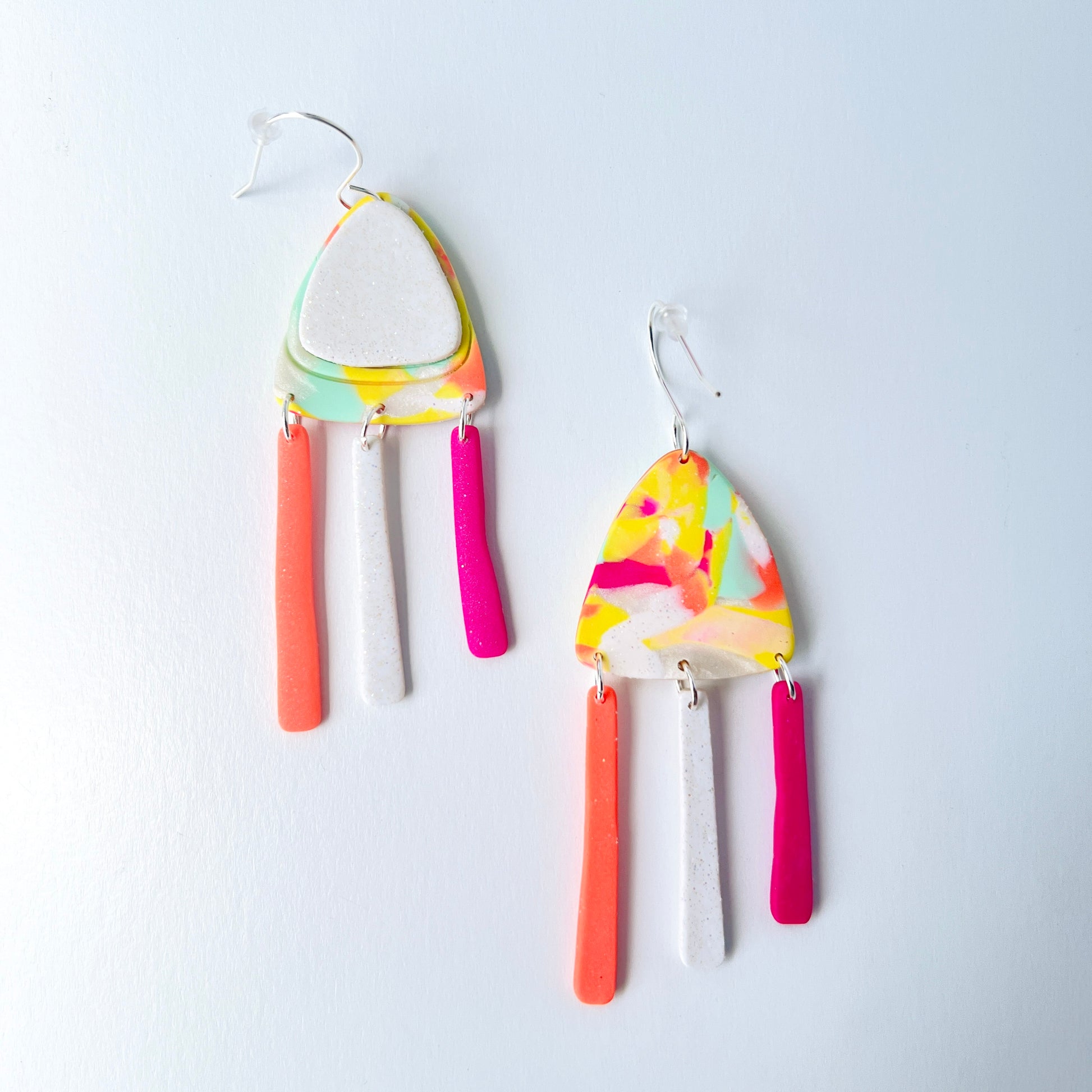 A pair of handmade polymer clay earrings with layered triangle shapes. The top layer is speckled white, sitting on a colorful marbled triangle of yellow, green, pink, and coral. Each earring features three dangles: one neon coral, one speckled white, and one bright pink, all hanging from silver stainless steel earwires.