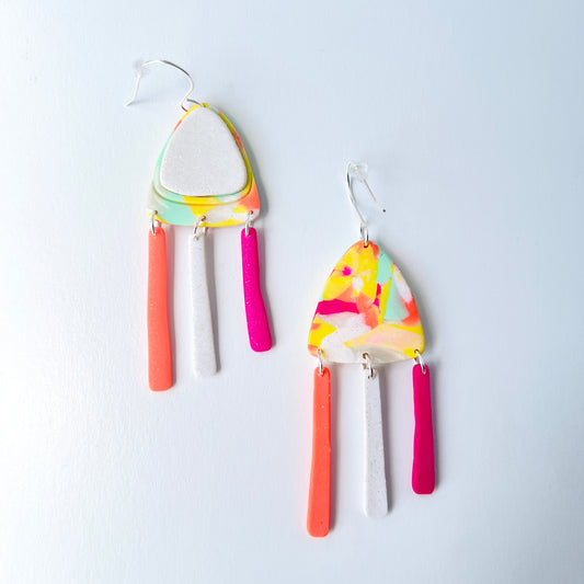 A pair of handmade polymer clay earrings with layered triangle shapes. The top layer is speckled white, sitting on a colorful marbled triangle of yellow, green, pink, and coral. Each earring features three dangles: one neon coral, one speckled white, and one bright pink, all hanging from silver stainless steel earwires.