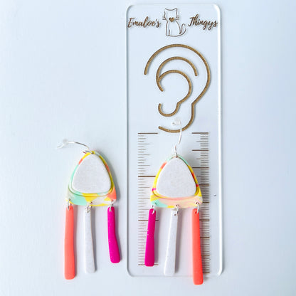 A pair of handmade polymer clay earrings with layered triangle shapes. The top layer is speckled white, sitting on a colorful marbled triangle of yellow, green, pink, and coral. Each earring features three dangles: one neon coral, one speckled white, and one bright pink, all hanging from silver stainless steel earwires.