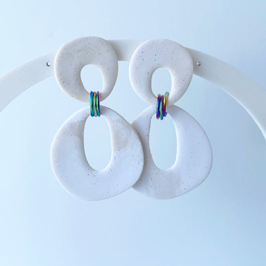 A pair of handmade dangle earrings crafted from shimmery white polymer clay in abstract looped shapes. Each earring features two interlocking hollow ovals connected by colorful rainbow-toned jump rings. Mounted on titanium posts, the earrings are lightweight and modern, photographed against a light background