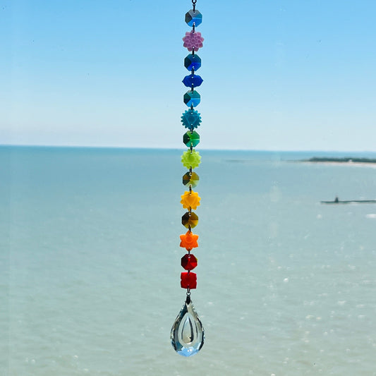 A handmade chakra suncatcher featuring seven stacked polymer clay beads in the colors of the chakras, strung vertically and finished with a hanging faceted glass crystal that reflects rainbows when hit by sunlight. Designed for spiritual, boho, or meditative decor spaces.