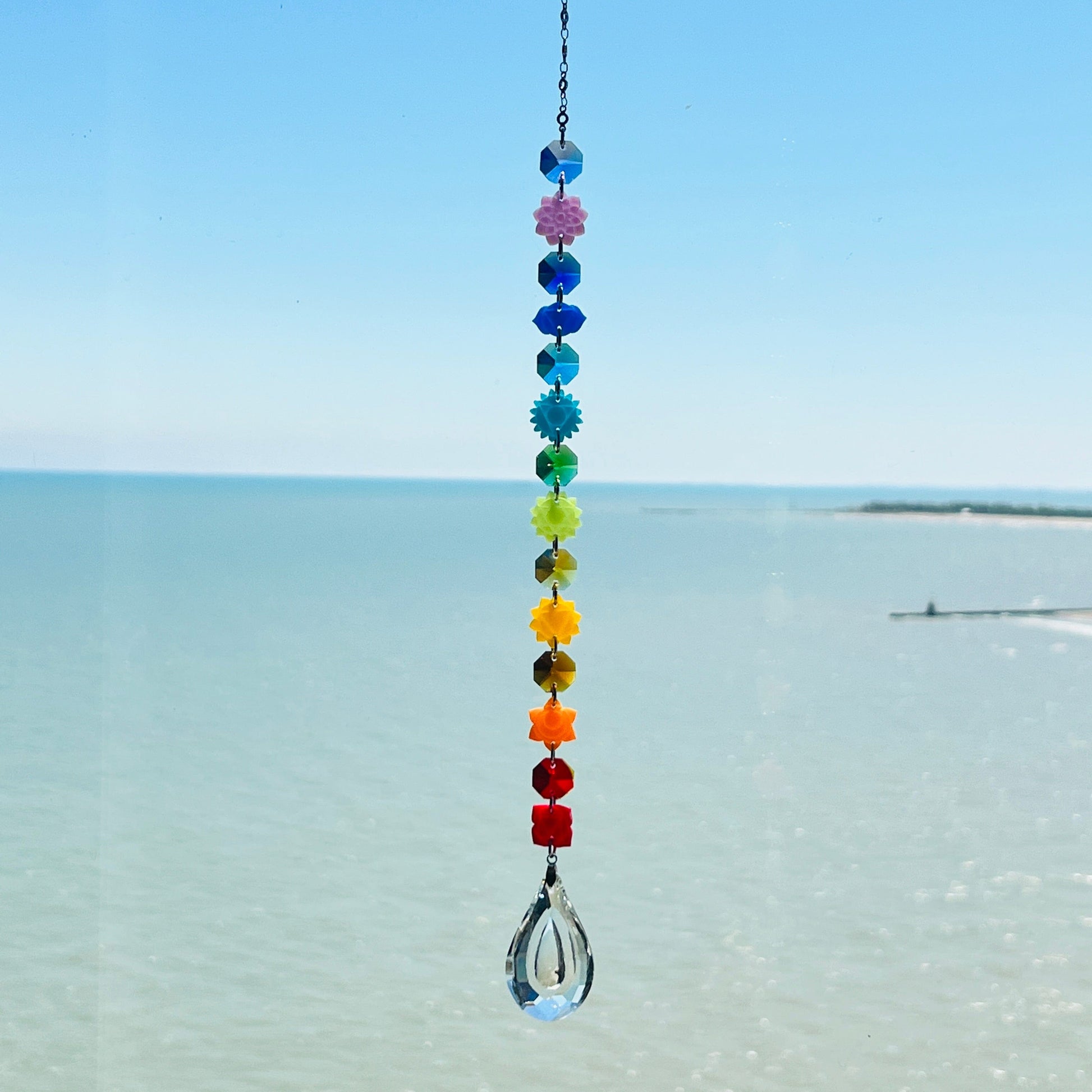 A handmade chakra suncatcher featuring seven stacked polymer clay beads in the colors of the chakras, strung vertically and finished with a hanging faceted glass crystal that reflects rainbows when hit by sunlight. Designed for spiritual, boho, or meditative decor spaces.