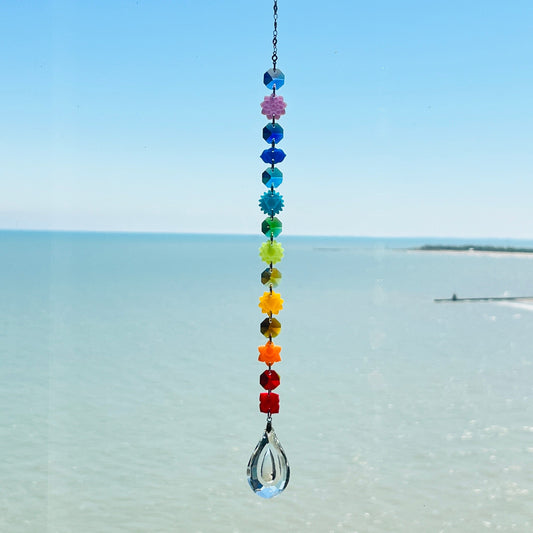 A handmade chakra suncatcher featuring seven stacked polymer clay beads in the colors of the chakras, strung vertically and finished with a hanging faceted glass crystal that reflects rainbows when hit by sunlight. Designed for spiritual, boho, or meditative decor spaces.