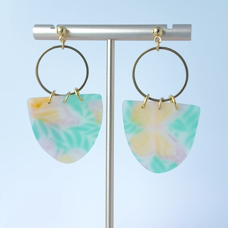 Video of pair of handmade earrings featuring semi-transparent polymer clay shields with soft pastel patterns of yellow butterflies and green leaves, attached to gold-plated hoops and ball posts.  
