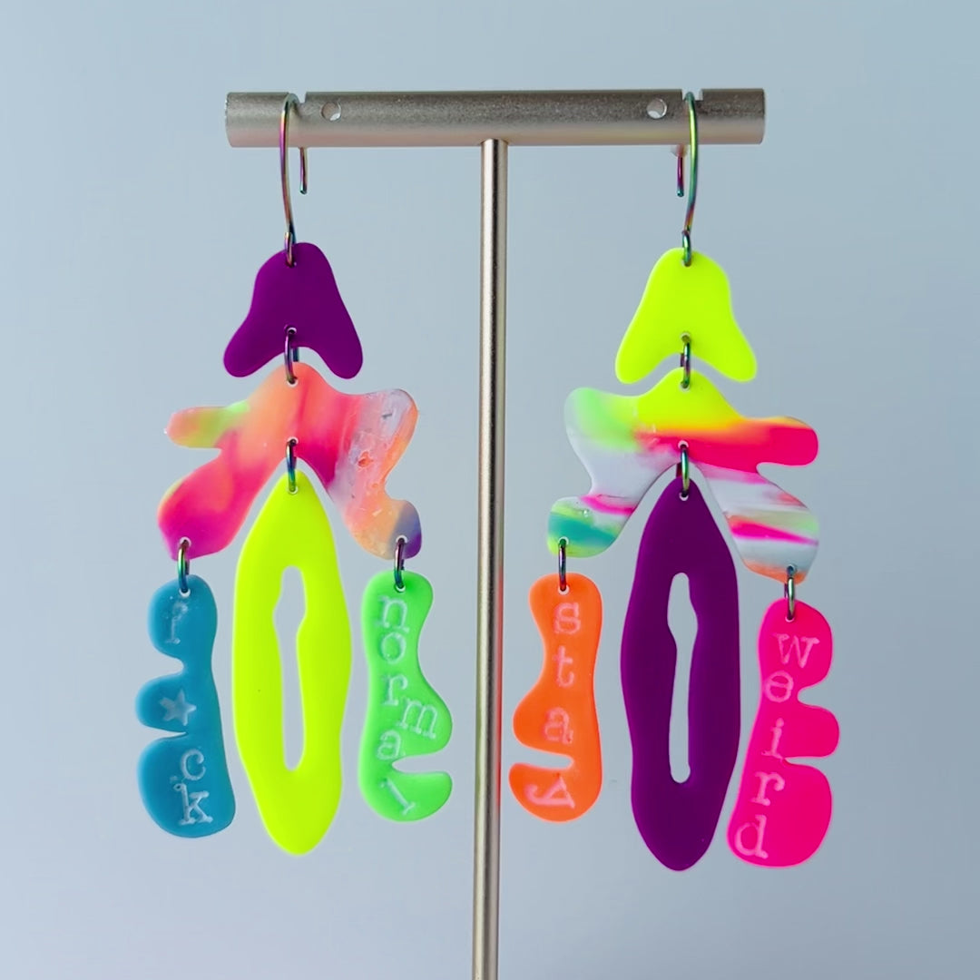 A video of pair of mismatched handmade statement earrings hanging from a gold-T bar spinning slowly in a circle.  The earrings are made from brightly colored neon polymer clay in layered abstract shapes. The left earring includes shapes in purple, orange, yellow, blue, and green and spells “F*ck Normal.” The right earring features neon pink, yellow, orange, purple, and green elements spelling “Stay Weird.” Both earrings are suspended from rainbow-plated stainless steel earwires