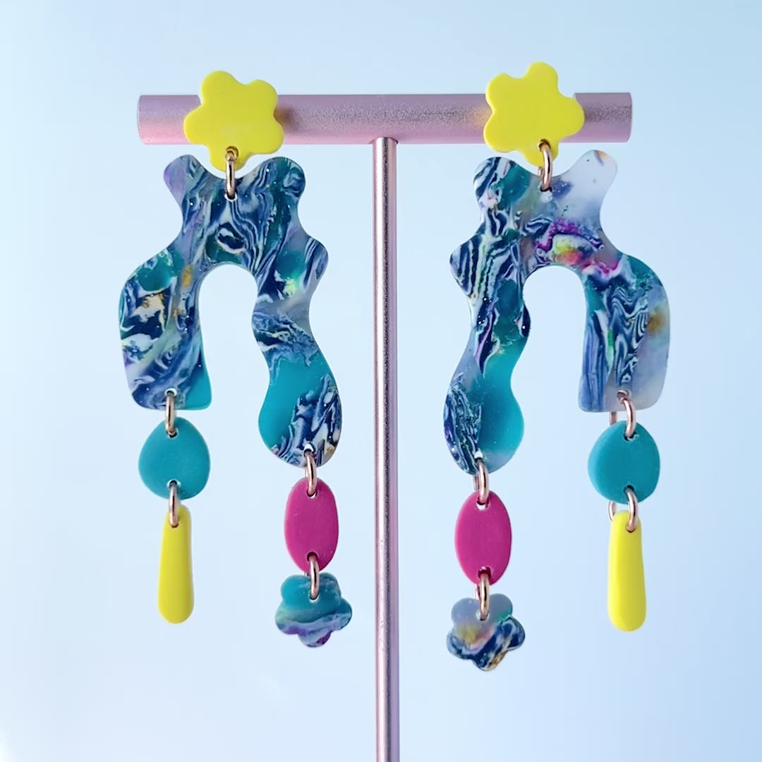 Video of a Colorful handmade polymer clay earrings featuring abstract, curvy marbled shapes in purple, blue, white, and pink with dangling components including teal and pink ovals, yellow drops, and blue flower accents. Topped with bright yellow flower studs and mounted on titanium posts