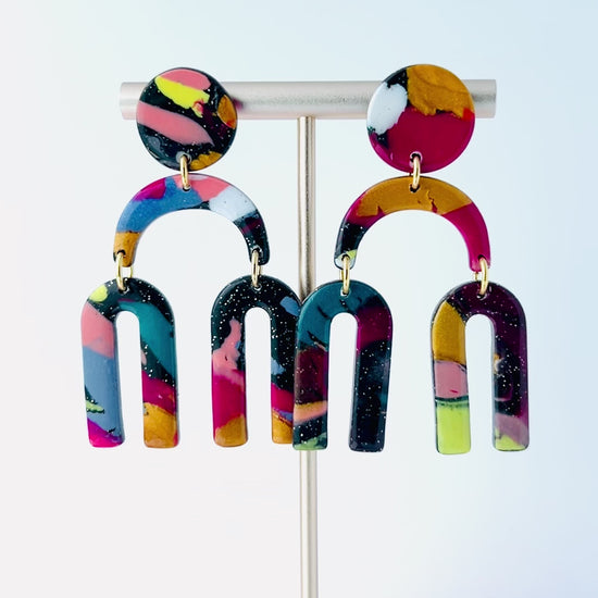 Video of a vibrant pair of handmade polymer clay dangle earrings shaped like arched rainbows with U-shaped dangles. Marbled in a mix of pink, teal, gold, white, black, and glitter, with titanium posts. Displayed against a white background