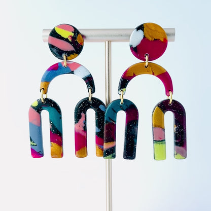Video of a vibrant pair of handmade polymer clay dangle earrings shaped like arched rainbows with U-shaped dangles. Marbled in a mix of pink, teal, gold, white, black, and glitter, with titanium posts. Displayed against a white background