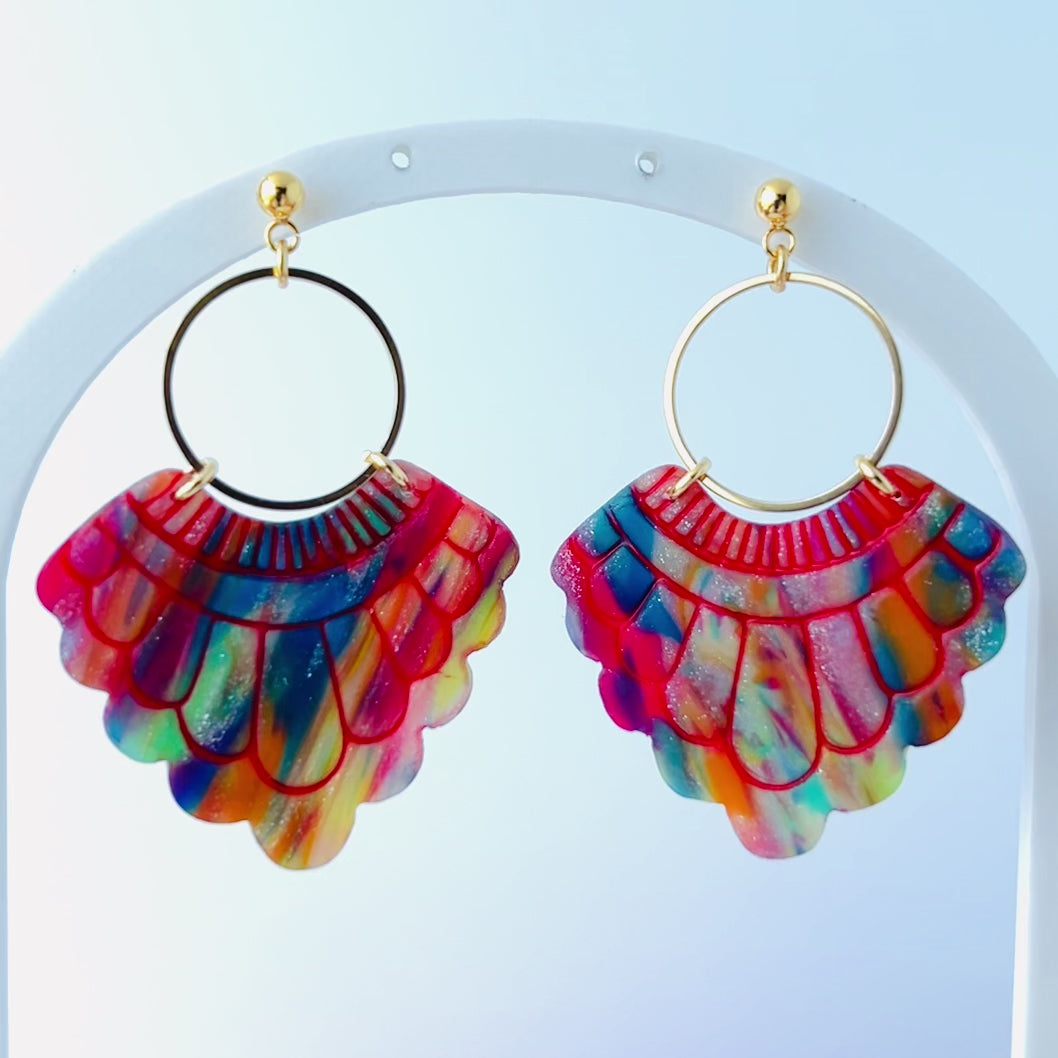 Video of a pair of handmade fan-shaped polymer clay earrings displayed on a white background. The earrings feature gold-plated ball posts and hoops, with semi-transparent rainbow-colored clay and red etched fan-like lines. The marbling effect shows swirls of pink, blue, green, orange, and white.