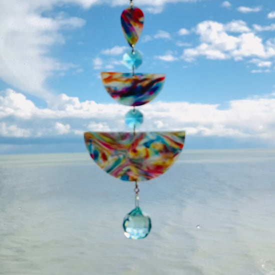 Video of Golden Swirl: Swirled warm-toned polymer clay teardrop and arches with aqua glass crystals and a gold chain, photographed against a bright sky over water.