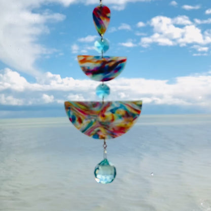 Video of Golden Swirl: Swirled warm-toned polymer clay teardrop and arches with aqua glass crystals and a gold chain, photographed against a bright sky over water.