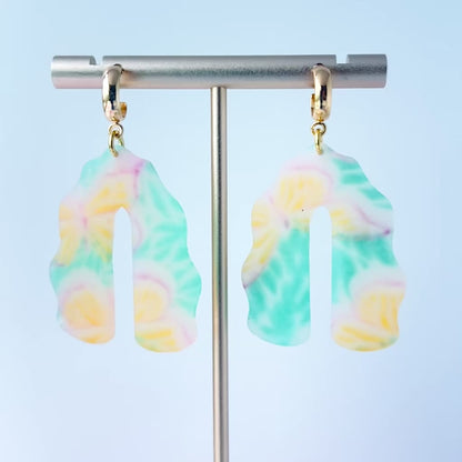 Video of a pair of handmade polymer clay earrings shaped like arches, with swirled pastel colors and subtle details of yellow butterflies and green leafy accents. The semi-transparent clay design is paired with shiny gold-plated brass half hoop posts.