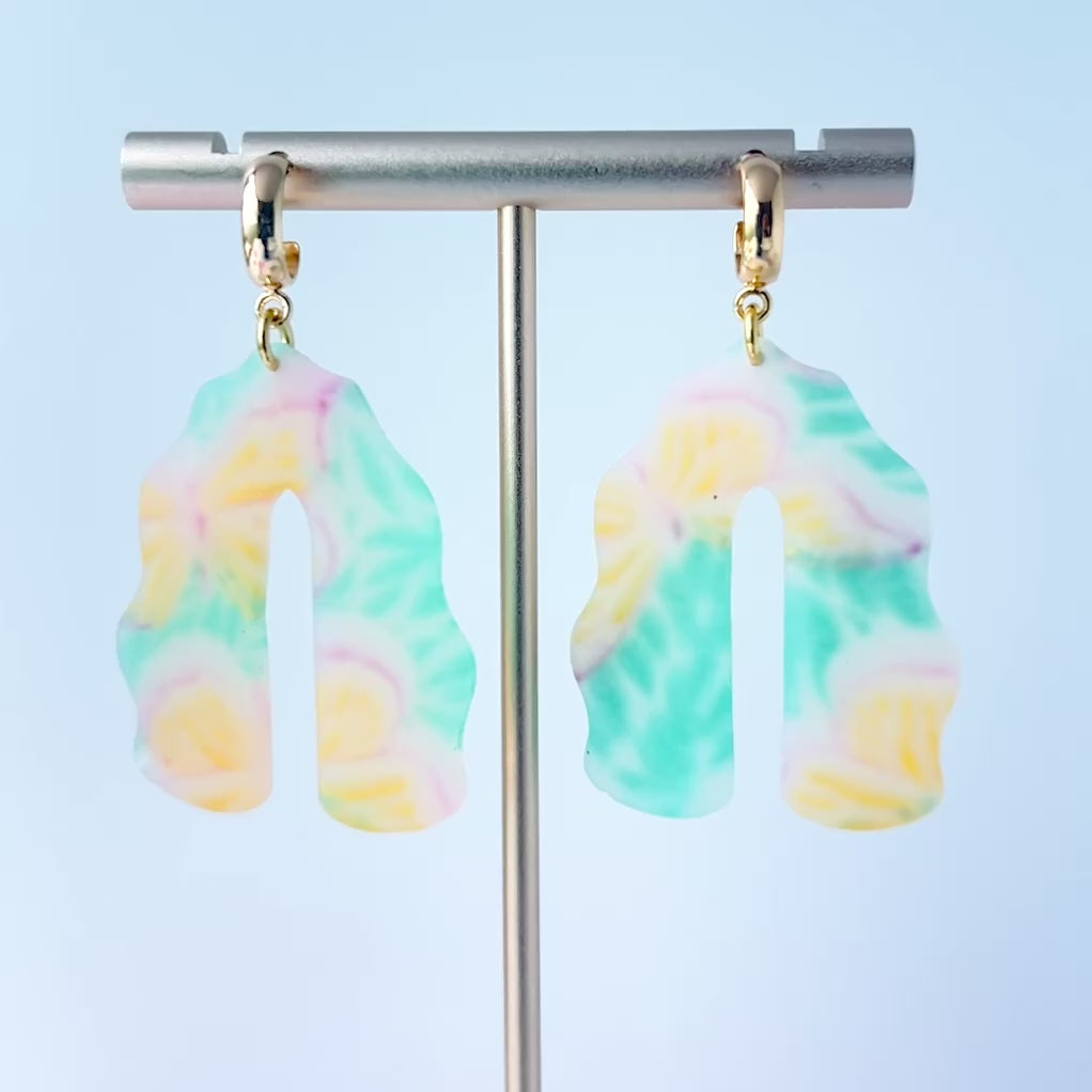 Video of a pair of handmade polymer clay earrings shaped like arches, with swirled pastel colors and subtle details of yellow butterflies and green leafy accents. The semi-transparent clay design is paired with shiny gold-plated brass half hoop posts.
