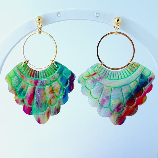 Video of a pair of handmade fan-shaped polymer clay earrings displayed on a white background. The earrings feature gold-plated ball posts and hoops, with semi-transparent rainbow-colored clay and green etched fan-like lines. The marbling effect shows swirls of pink, blue, green, orange, and white.