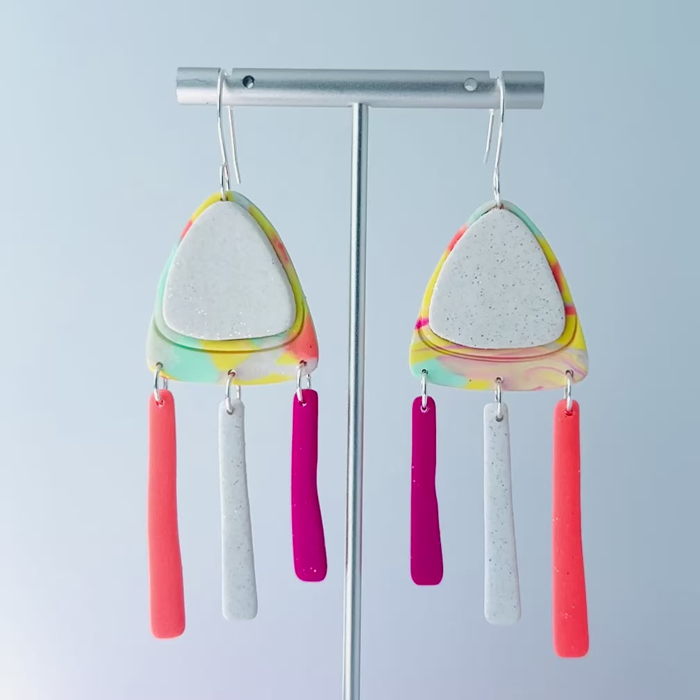 Video of a pair of handmade polymer clay earrings with layered triangle shapes. The top layer is speckled white, sitting on a colorful marbled triangle of yellow, green, pink, and coral. Each earring features three dangles: one neon coral, one speckled white, and one bright pink, all hanging from silver stainless steel earwires.