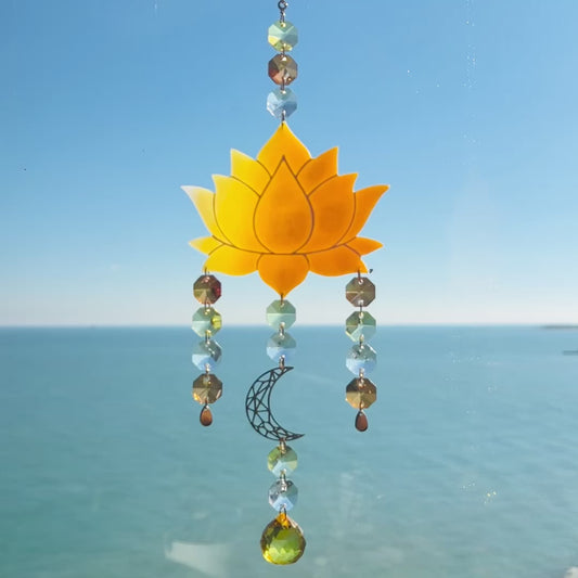 Video of a handmade lotus flower suncatcher crafted from ombre orange & yellow polymer clay, featuring a stylized lotus bloom suspended from a gold chain with a faceted glass crystal hanging below, designed to reflect rainbow light in the sun. Ideal for meditation rooms, spiritual decor, or peaceful window displays.