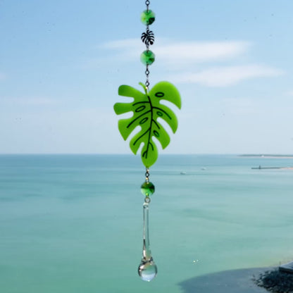 Video of a handmade monstera leaf suncatcher made from green polymer clay with detailed vein textures, hanging from a gold loop and chain, featuring a faceted glass crystal that casts rainbows in sunlight. Ideal for botanical or boho home decor.