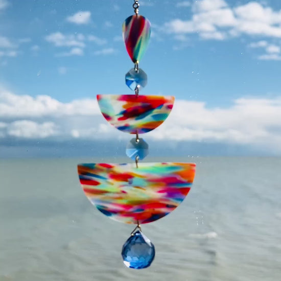 video of Paint Palette: Colorful handmade suncatcher with bold primary-toned polymer clay pieces in teardrop and arch shapes, paired with translucent blue faceted crystals and a rainbow chain.