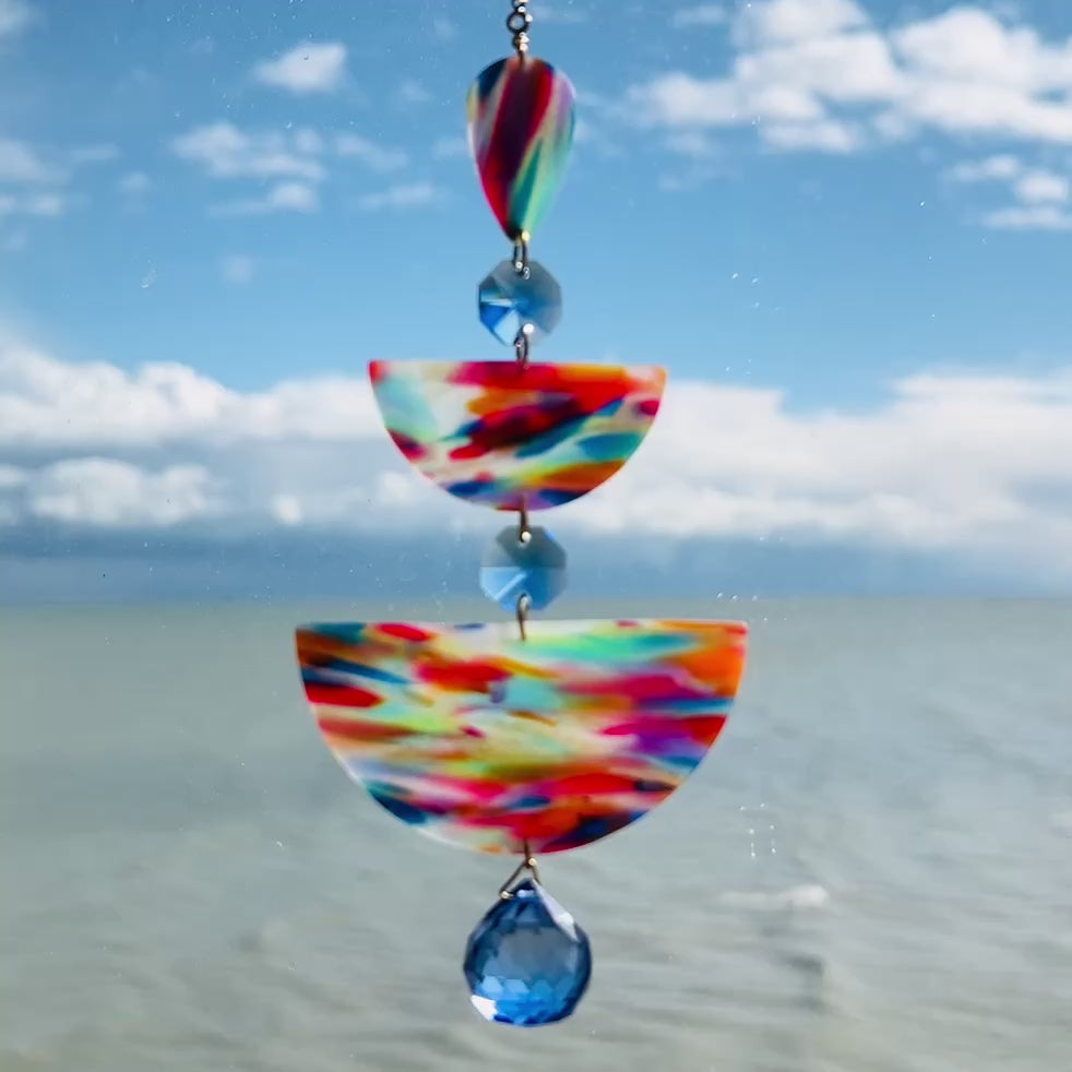 video of Paint Palette: Colorful handmade suncatcher with bold primary-toned polymer clay pieces in teardrop and arch shapes, paired with translucent blue faceted crystals and a rainbow chain.
