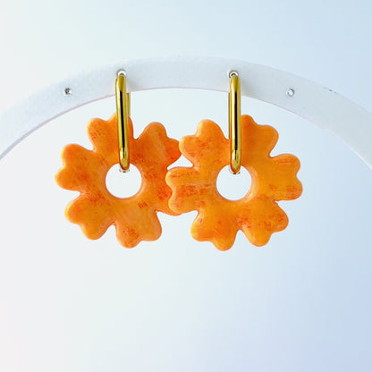 Video of a pair of orange handmade polymer clay earrings shaped like abstract flowers, each dangling from a gold oval-shaped 316L stainless steel hoop. 