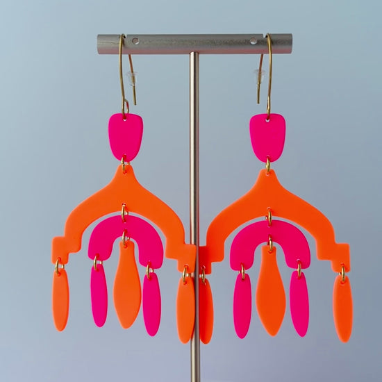 Video of a pair of vibrant handmade chandelier earrings with hot pink and neon orange polymer clay components, featuring multiple dangling shapes on gold-plated stainless steel earwires.