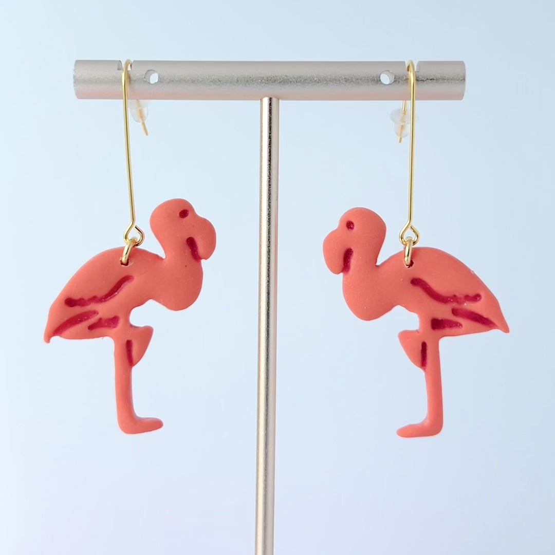 Video of a pair of handmade coral-pink flamingo earrings sculpted from polymer clay, each suspended from long, minimalist gold-plated stainless steel bar earwires. The flamingos face each other in a mirrored design.  The earrings are on a slow spinning display showing the earrings from all sides
