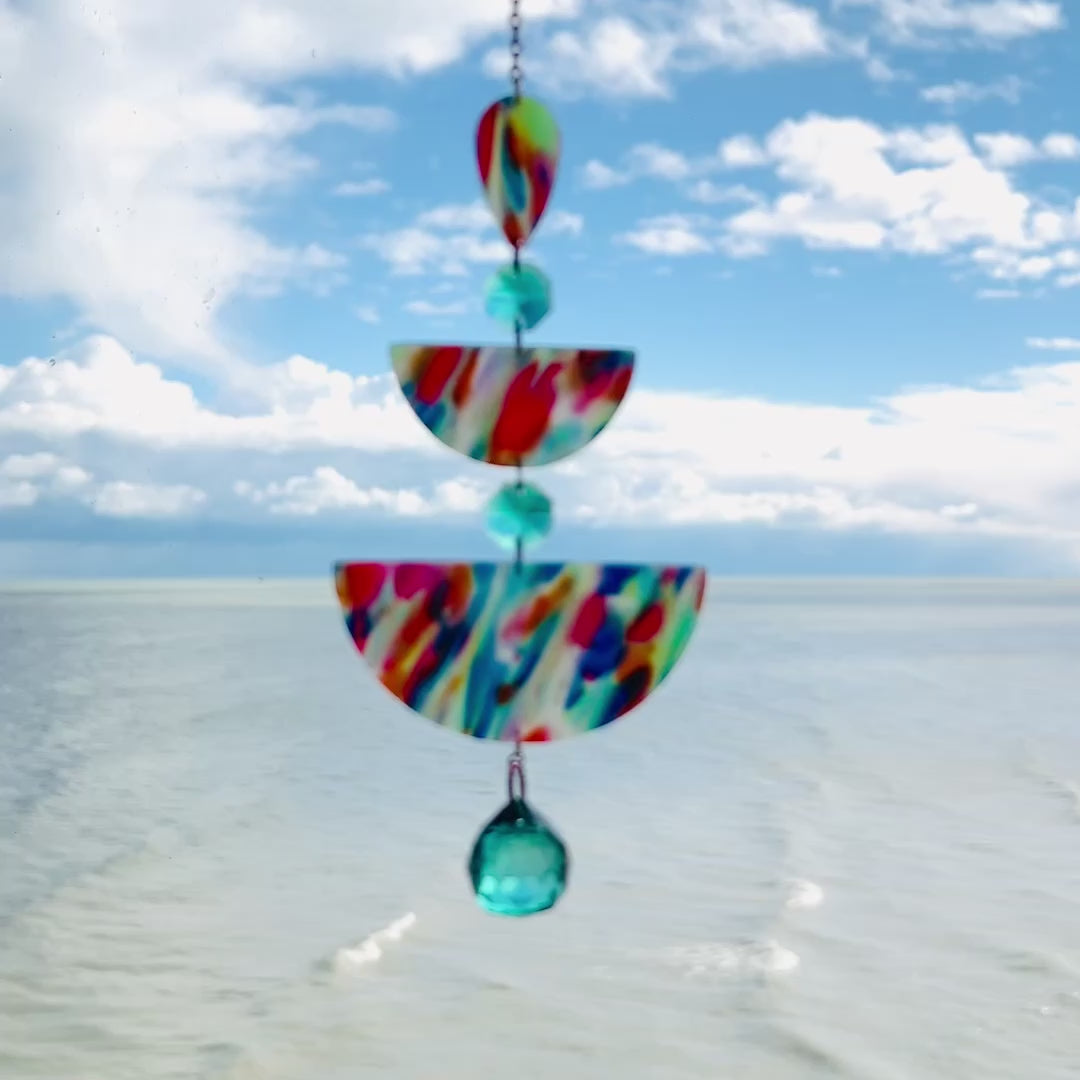 Video of Rainbow Rain: A handmade suncatcher hanging in front of a blue sky, featuring marbled rainbow polymer clay shapes in teardrop and half-circle forms, accented with sea green glass crystals and a rainbow chain.