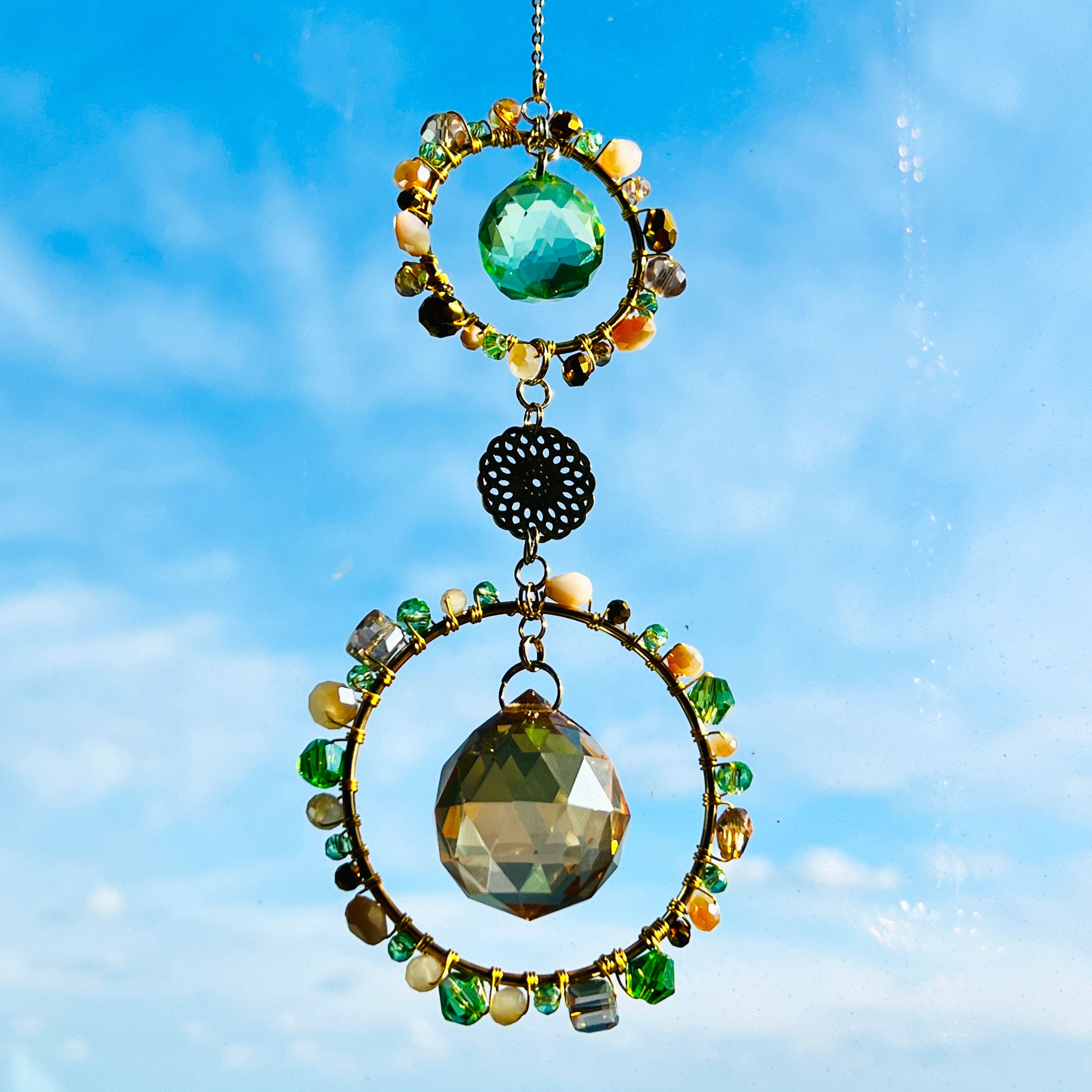 A handmade suncatcher with two gold wire-wrapped rings adorned with green, gold, pearl, and iridescent glass beads. A smaller ring at the top features a green faceted crystal, while the larger bottom ring features a champagne-gold crystal prism. Both are connected by a decorative round gold filigree charm and hang from a gold chain, photographed against a bright blue sky.